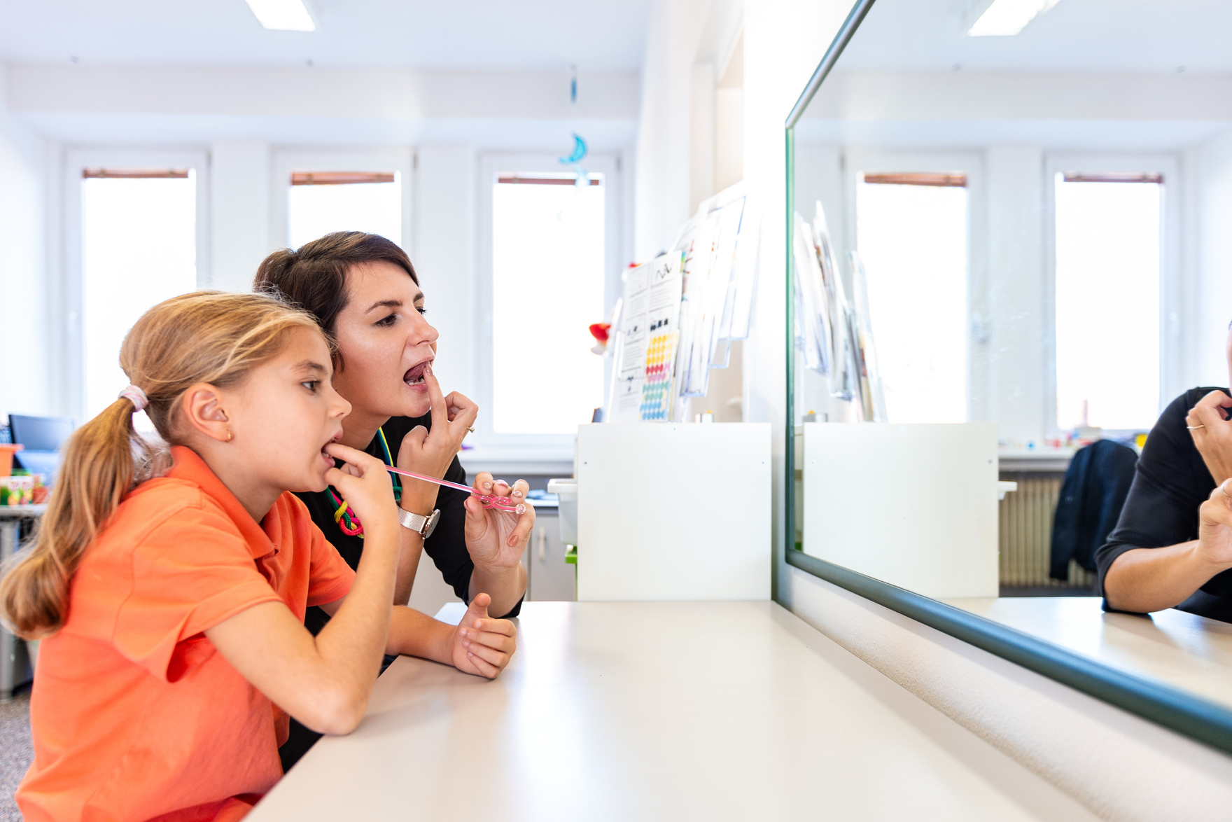 Young girl in speech therapy office. Mirror reflection of young girl exercising correct pronunciation with speech therapist.